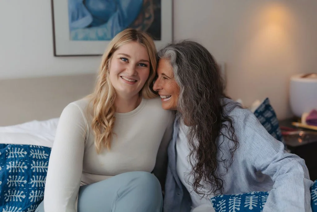A young woman and an older woman sitting on a bed, smiling at each other in a warmly lit room.