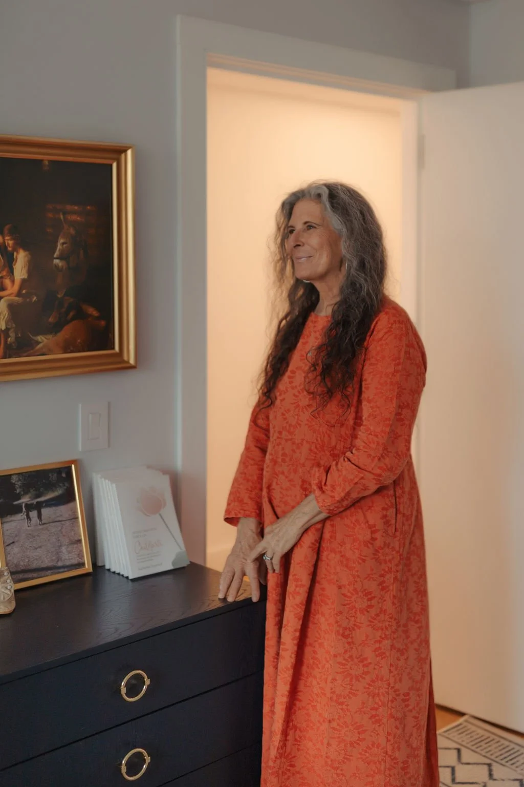A woman with long curly gray hair, wearing an orange patterned dress, standing next to a black dresser in a room with framed artwork and photographs on the wall and a stack of white paper or card. She is smiling and looking to the side.