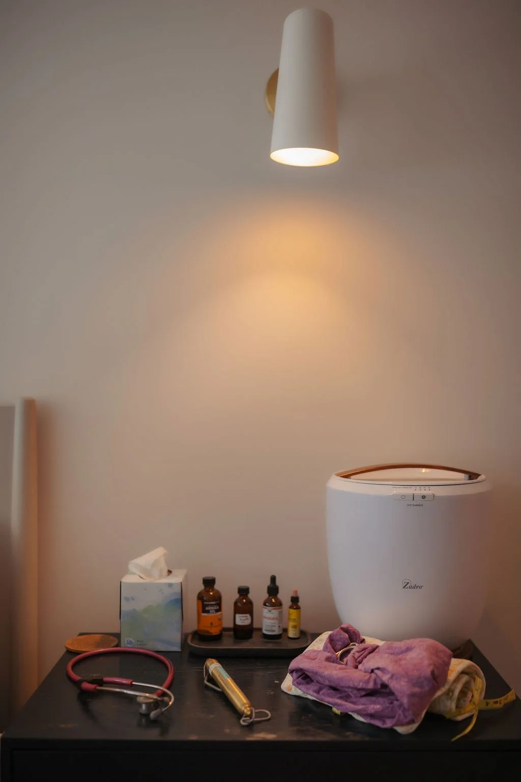 Medical supplies on a black table including a stethoscope, various medicine bottles, a tissue box, a gold medical tool, and some folded cloths, with a white medical device and a wall-mounted lamp above.