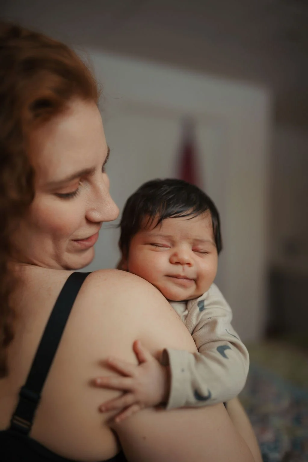 A woman holding a sleeping baby close to her shoulder, both with gentle, affectionate expressions.