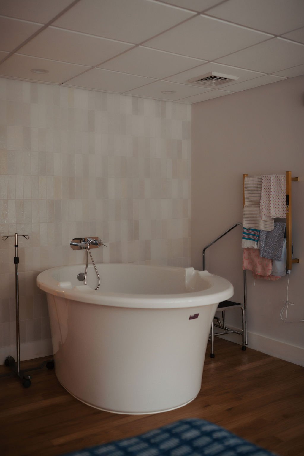 A medical bathing room with a white freestanding bathtub, an IV pole, and a towel rack with colorful towels on the wall. The room has beige tiled walls and a wooden floor.