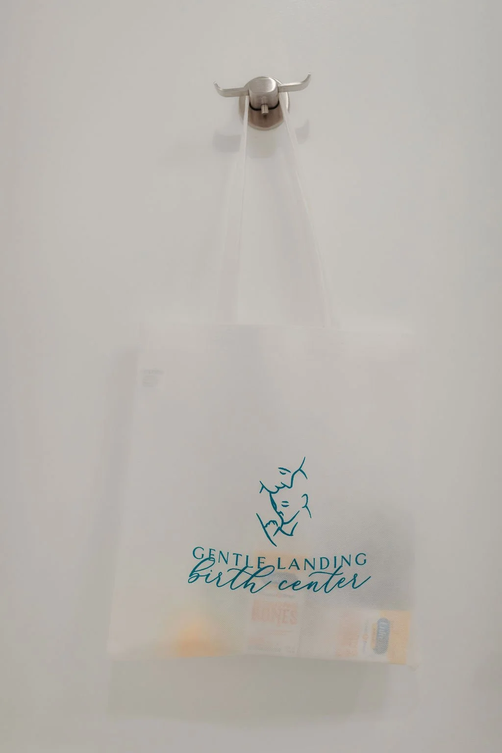 A paper bag hanging from a metal hook on the wall, with blue logo and text reading 'Gentle Landing Birth Center' and an outline sketch of a mother cradling her baby.