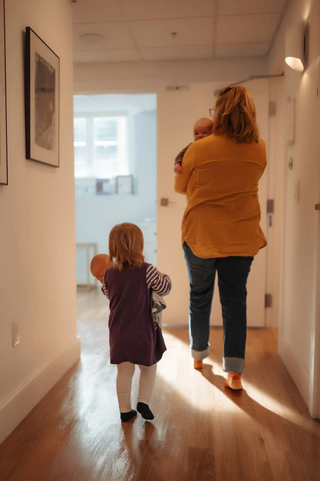 A woman walking down a hallway with two young children, one of whom is being carried and the other walking alongside. The hallway has wooden floors, framed pictures on the wall, and is lit by ceiling lights.