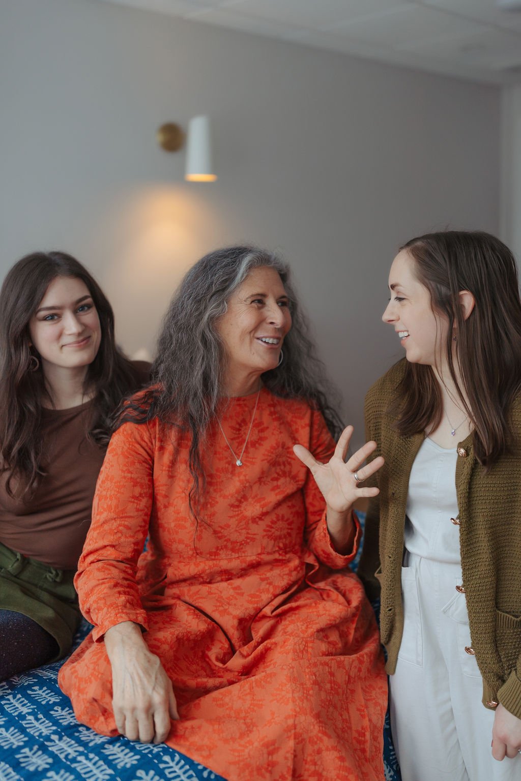Three women engaging in a lively conversation, with two of them smiling and one woman in the middle gesturing with her hand. They are indoors with a plain wall and wall lamp in the background.