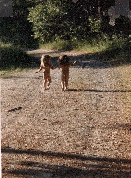 Two young children walking hand in hand on a dirt path in a wooded area during daytime.