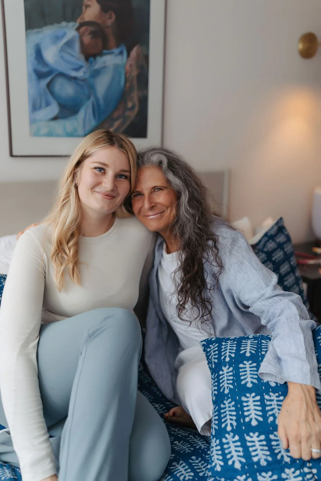 Two women, a young woman with blonde hair and an older woman with gray hair, sitting closely together on a patterned blue couch in a cozy room, smiling at the camera. A framed painting of a person holding a child hangs on the wall behind them.