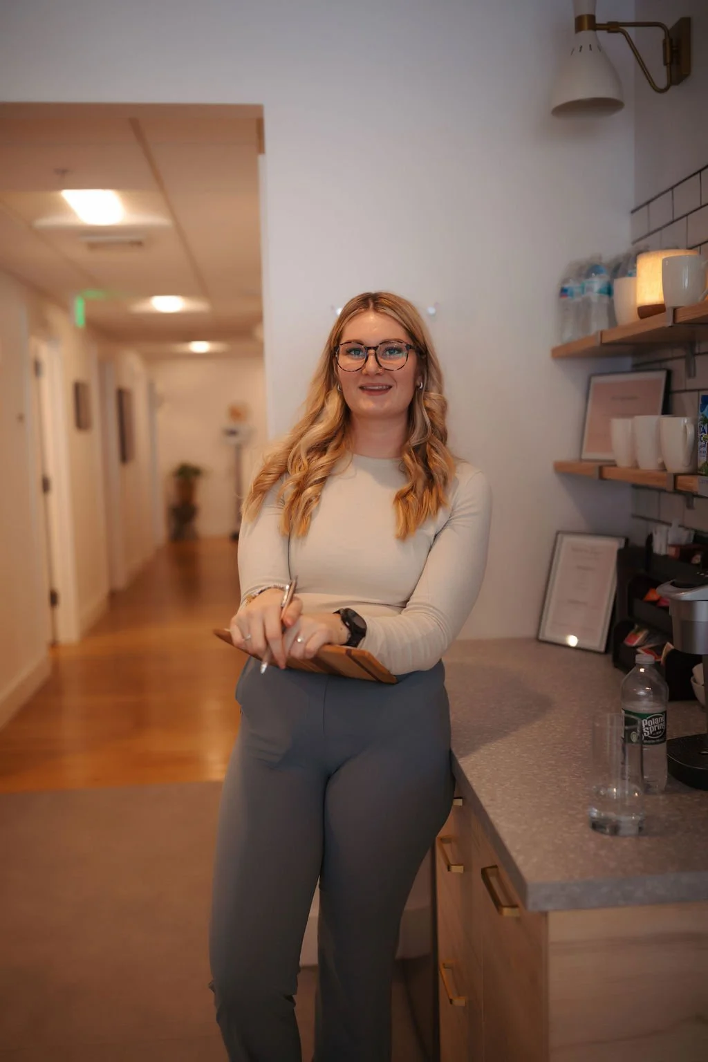 A woman with long blonde hair, glasses, wearing a light-colored long sleeve shirt and gray pants, standing in a modern room with wooden flooring and shelves, smiling at the camera.