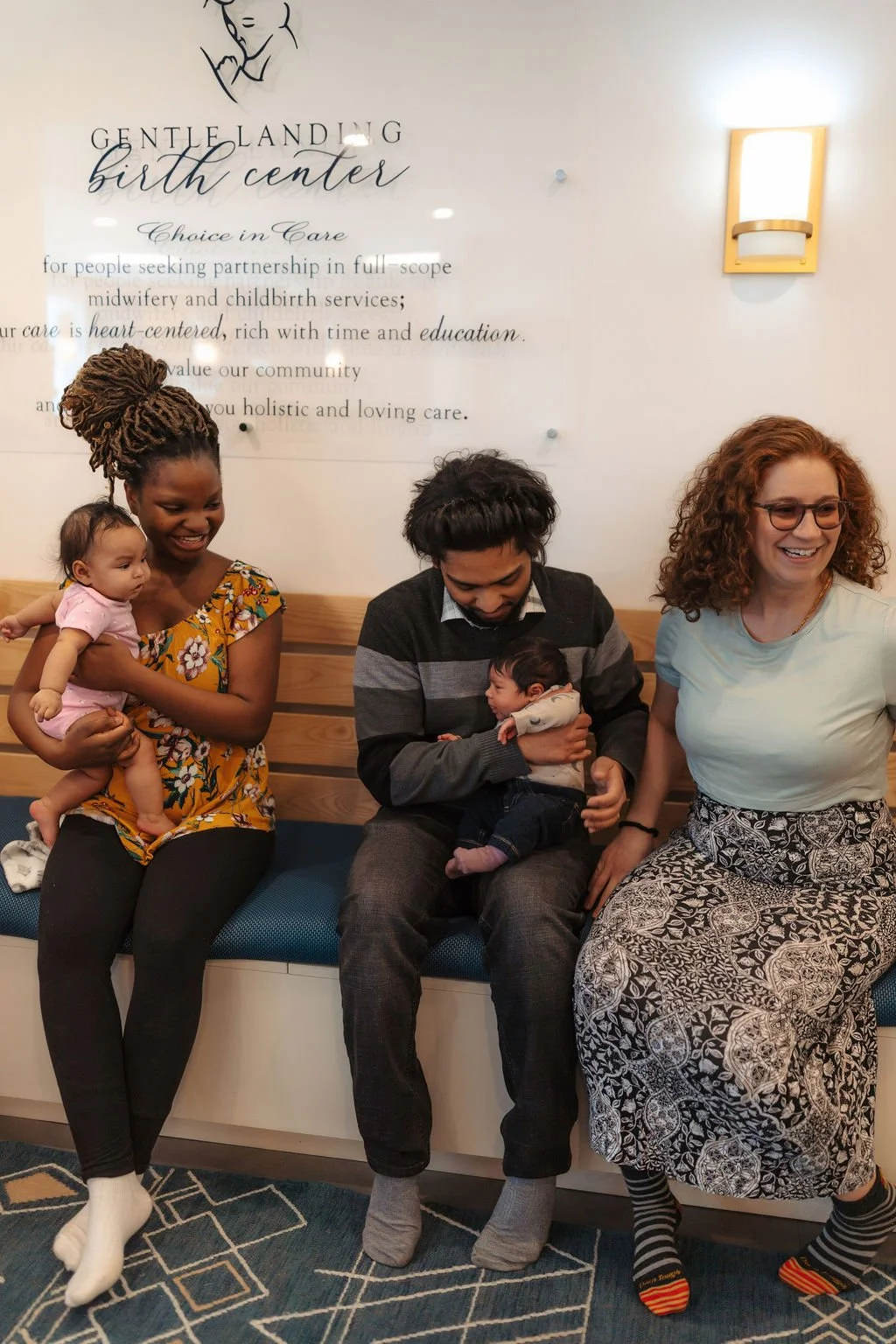 Group of four people, including two women and two men, sitting on a bench in a birth center, holding babies. The woman on the left has dreadlocks and is holding a small girl in pink. The man in the middle is looking at a newborn. The woman on the rig