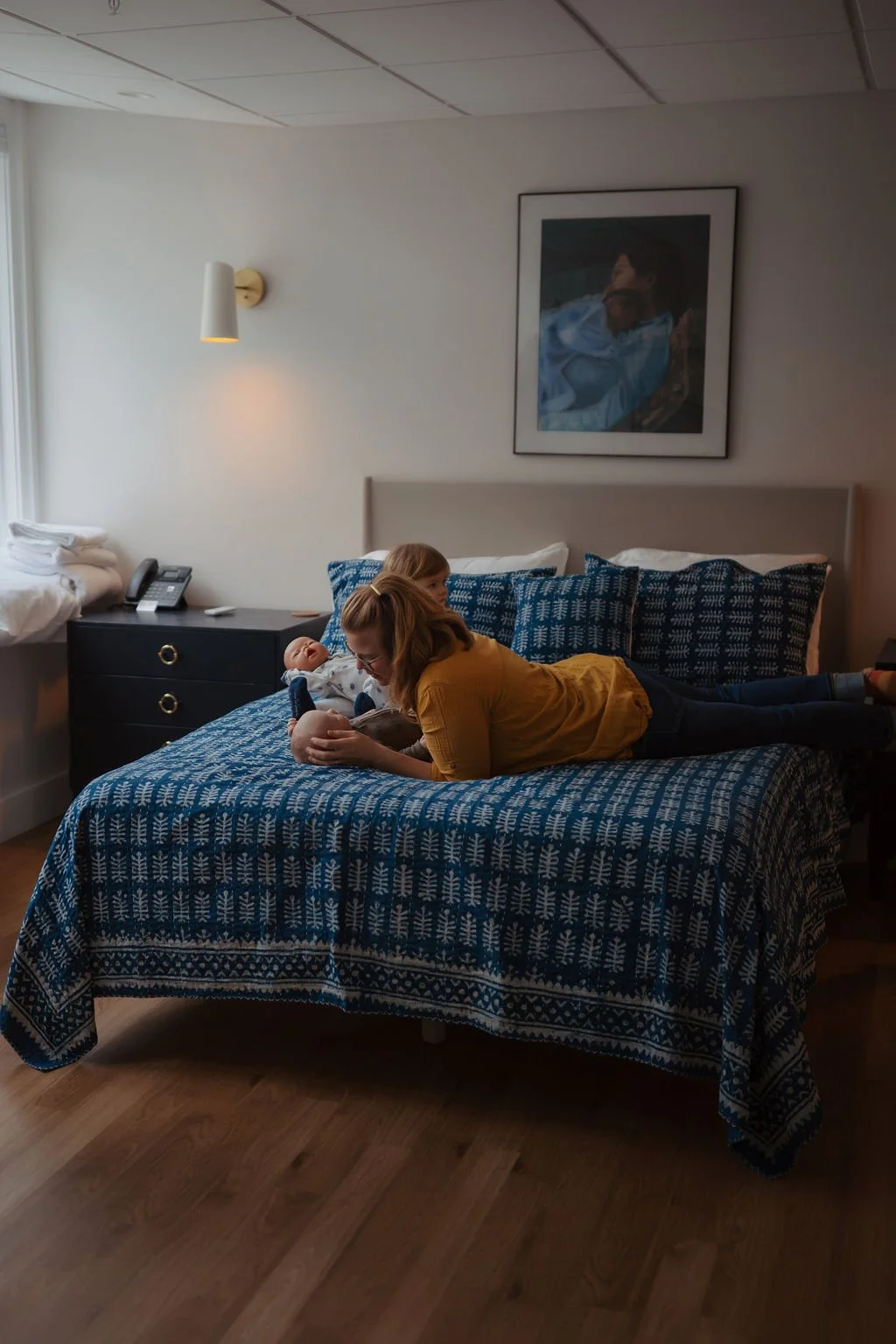 A woman is lying on her stomach on a bed, interacting with a baby who is lying on the bed. The woman has blonde hair, glasses, and is wearing a yellow shirt. The baby is dressed in a white outfit and is holding a stuffed toy. The bed has a blue patte