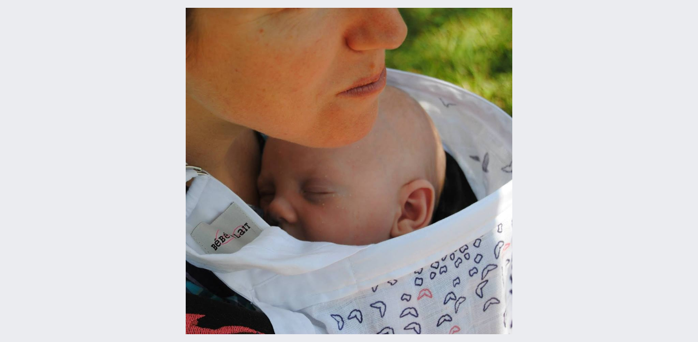 Close-up of a woman holding a sleeping baby in a white carrier outdoors.