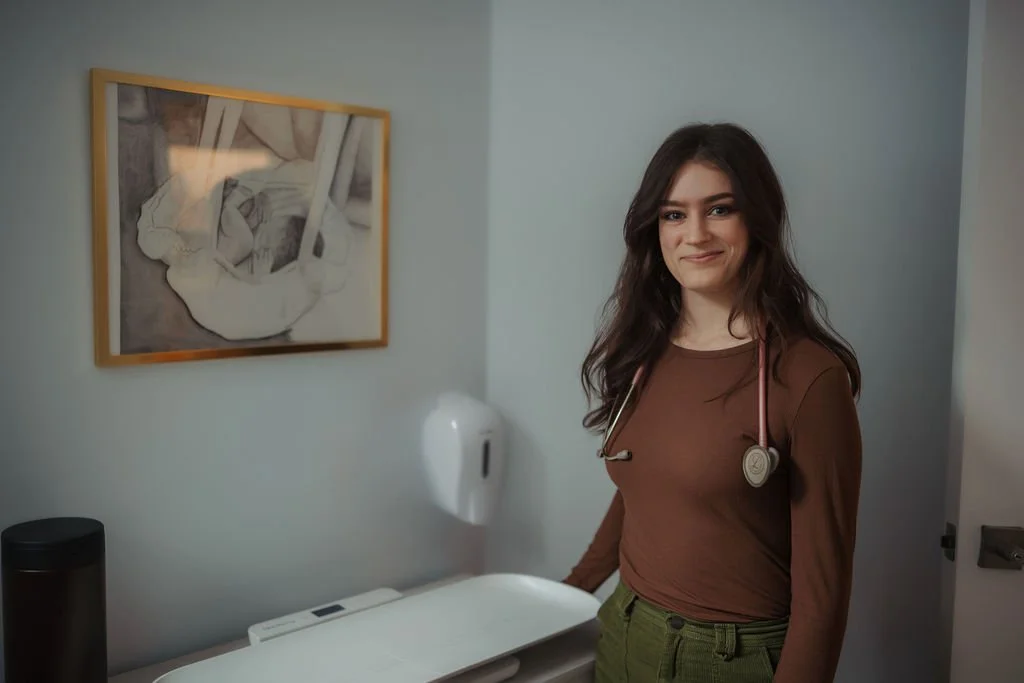A female doctor with a stethoscope around her neck standing beside an examination table in a clinic room.