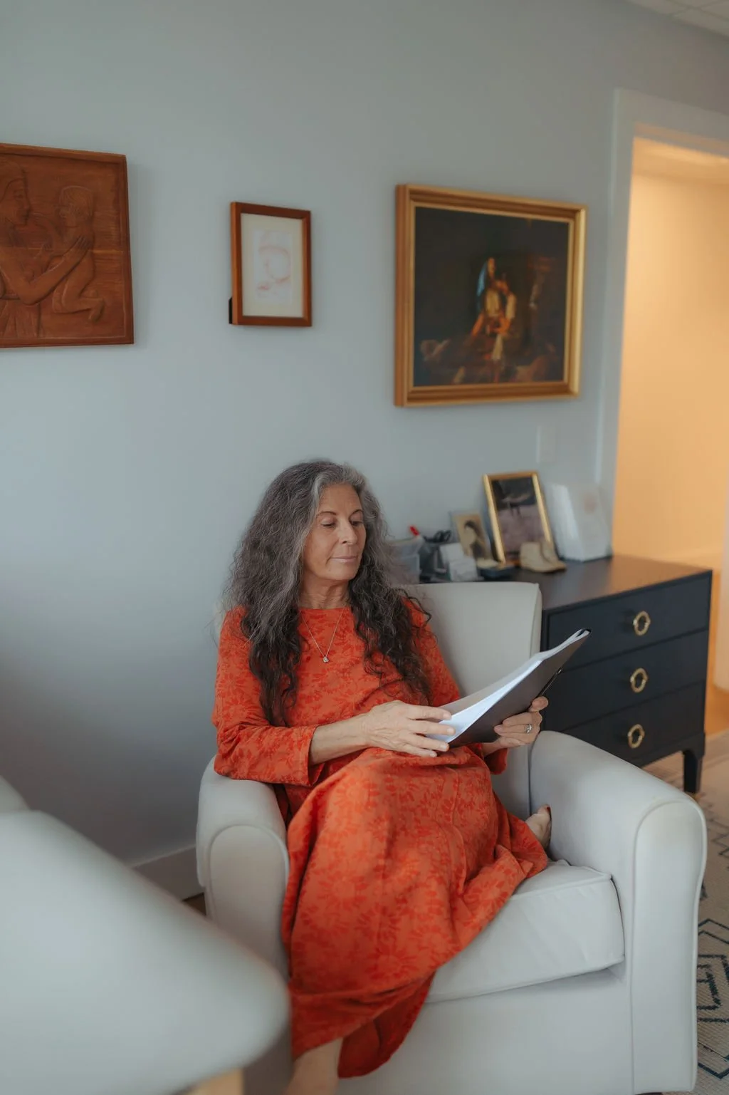 A woman with long gray hair wearing an orange dress sits in a white armchair, reading a notebook in a living room decorated with framed artwork on the wall and a black sideboard.