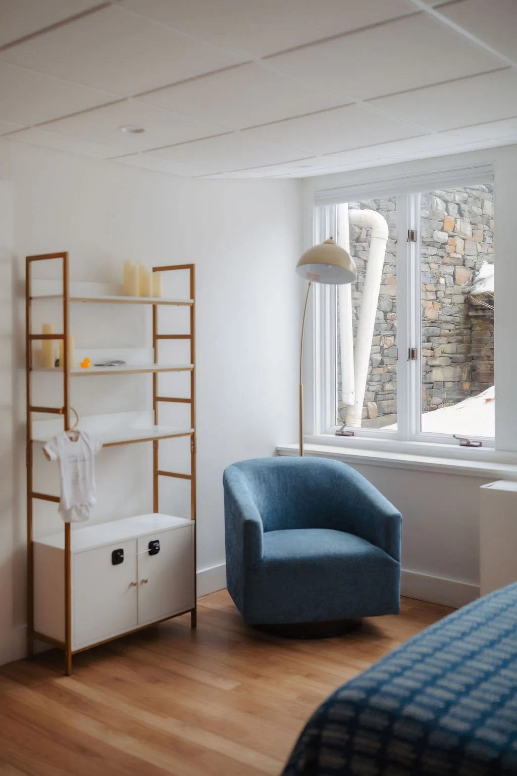 A cozy corner of a room with a blue armchair, a tall white standing lamp, a wooden and white shelving unit, and a window showing rocks and snow outside.