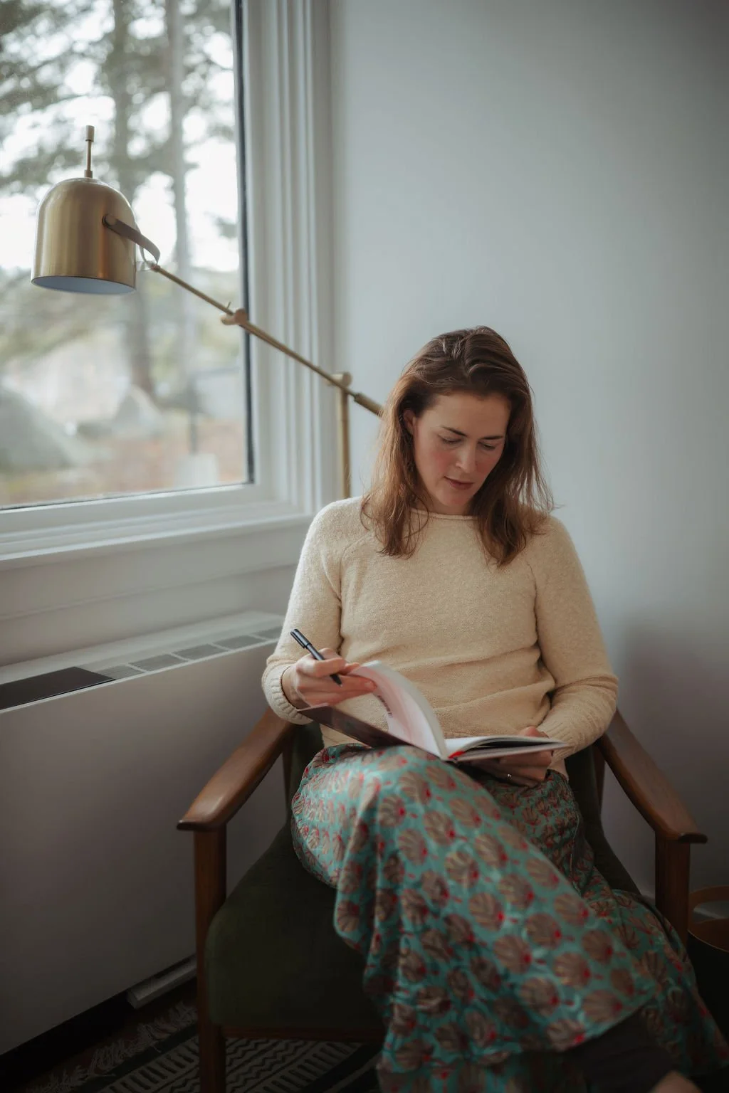 A woman sitting in a wooden chair, reading a book, with a pen in hand in a room near a window with white curtains and a brass wall-mounted lamp.