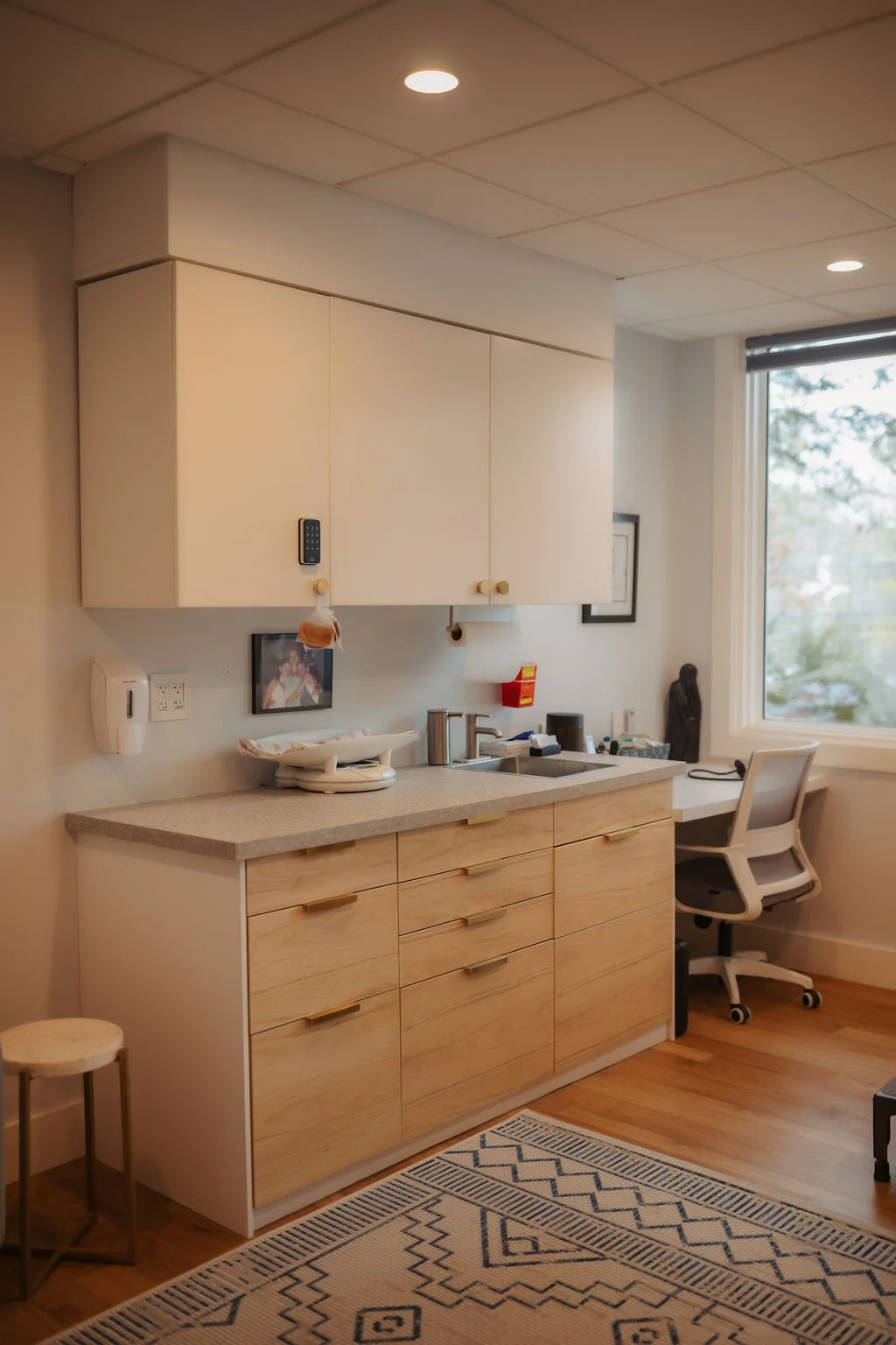Kitchen with white upper cabinets, wooden lower cabinets, a countertop with a sink, office chair near a window, and a patterned rug.