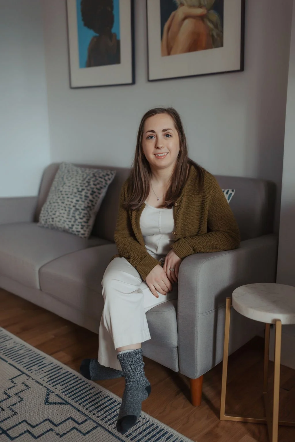 A woman with light skin and long brown hair, sitting on a light gray sofa in a living room, smiling at the camera. She is wearing a white top, brown cardigan, white pants, and gray socks. Behind her are two framed portraits on the wall, and a pattern