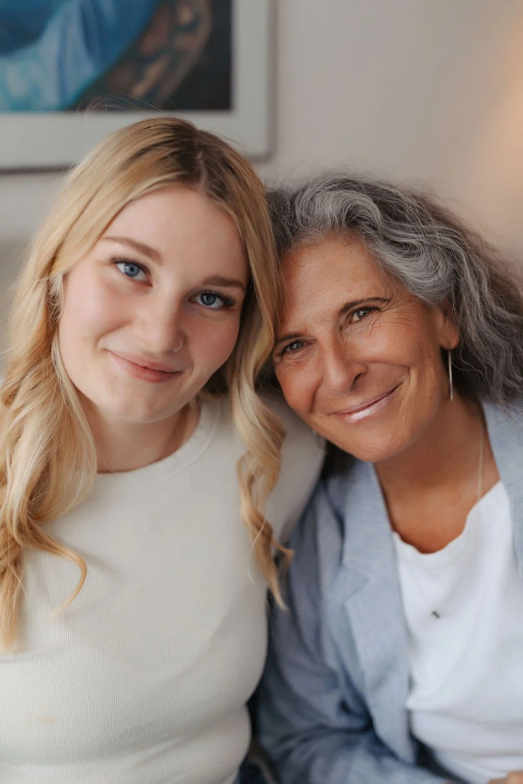A young woman with blonde hair and an older woman with gray curly hair smiling for a selfie indoors.