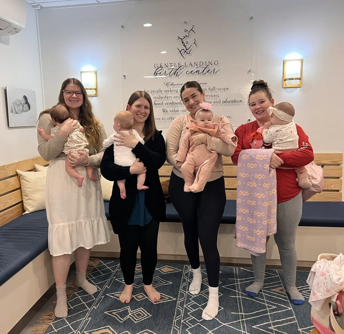 Four women holding babies inside a birth center waiting area with a sign that says 'Geneva Landing Birth Center' on the wall behind them.