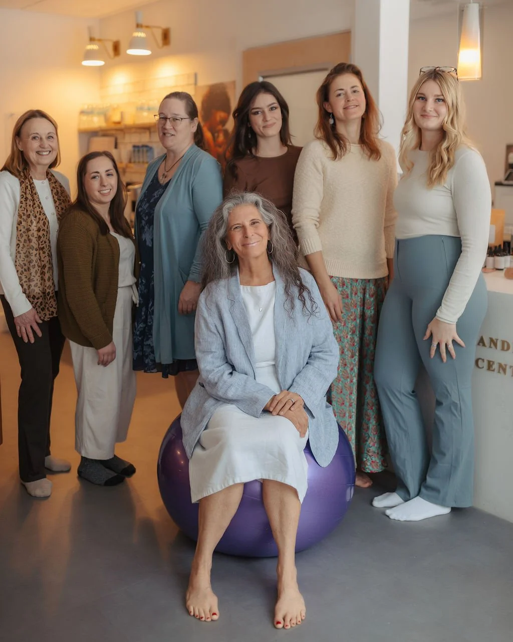 Group of women standing indoors, with one woman sitting on a purple exercise ball, smiling.