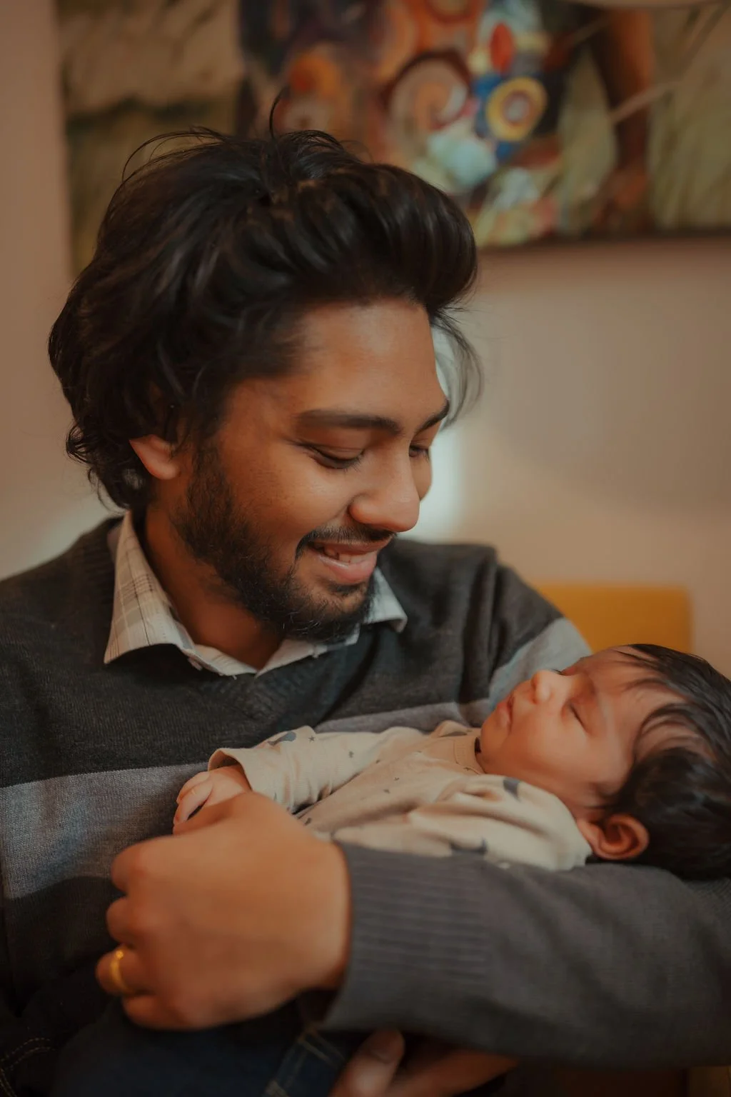 A man smiling warmly while holding a baby in his arms indoors.