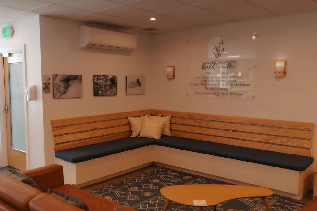 A waiting room with a wooden L-shaped bench with black cushions and beige pillows, a round wooden coffee table, framed photos on the wall, and a sign for a birth center.