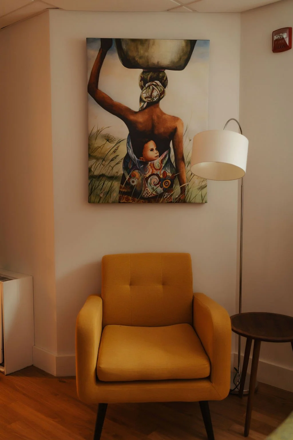 A yellow armchair with a small round side table and a floor lamp on a wooden floor in front of a wall with a large colorful painting of a woman carrying a pot on her head and a child resting on her back, with tall grass in the background.