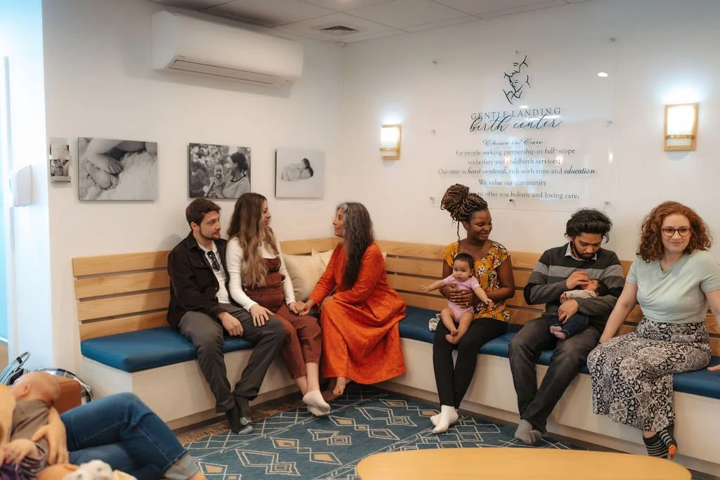 Group of people sitting in a waiting room or lounge area, including adults and children, with a wall sign indicating it is a childbirth center.