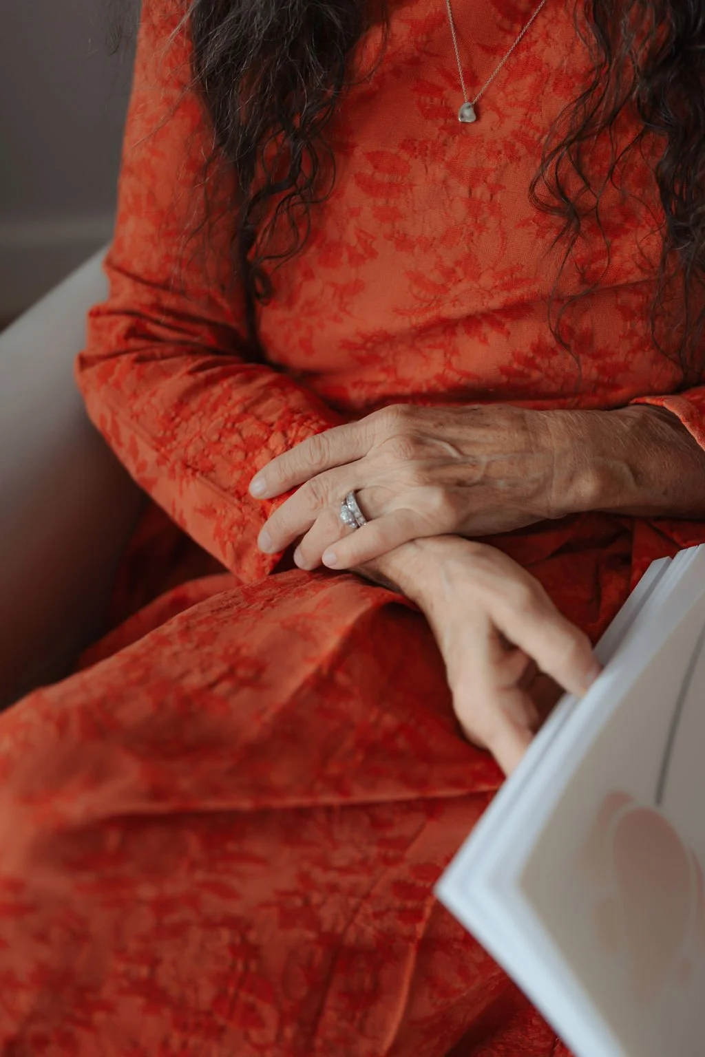 Close-up of a woman wearing a red patterned dress with a silver necklace, with her hands resting on her lap showing a wedding ring, sitting on a beige chair.