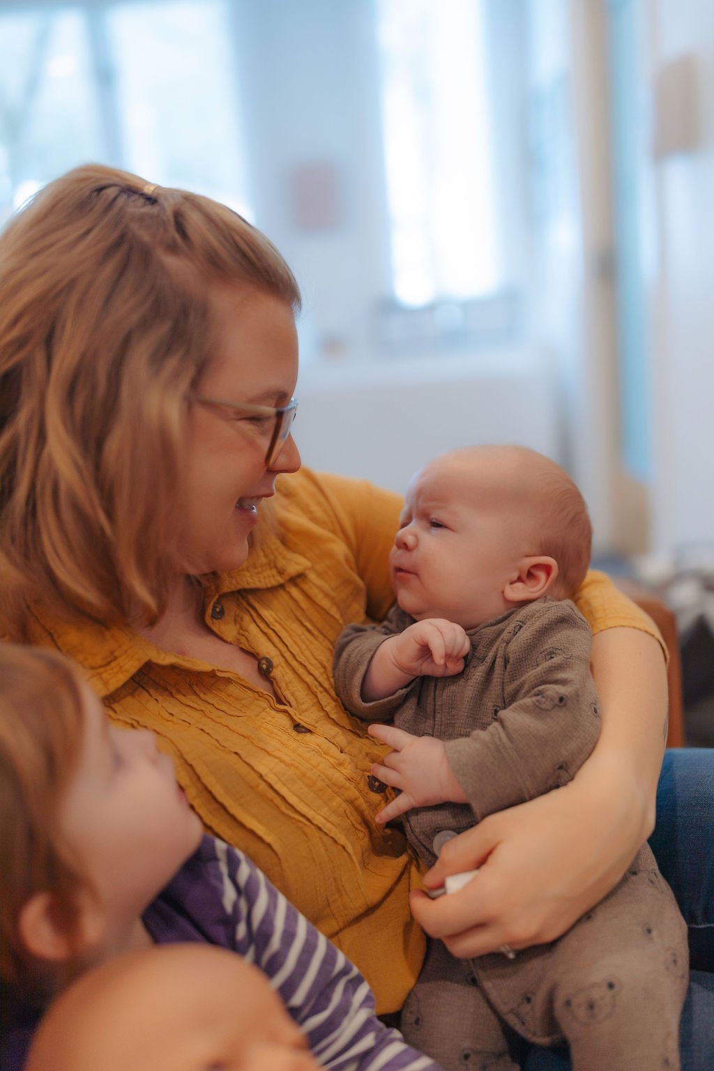 A woman with glasses and a mustard-colored shirt holding an infant, smiling at each other. A child with striped shirt is in the foreground.