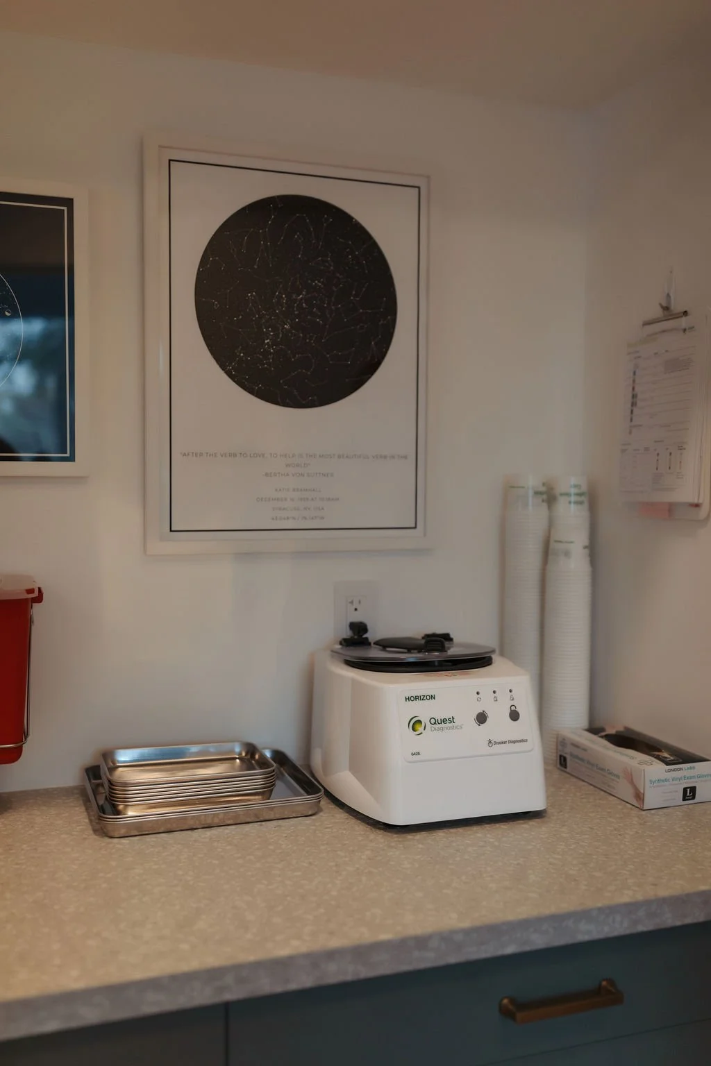 A countertop with a stack of metal trays, a medical instrument sterilizer, and two white standing tubes against a white wall with framed star maps and informational posters.