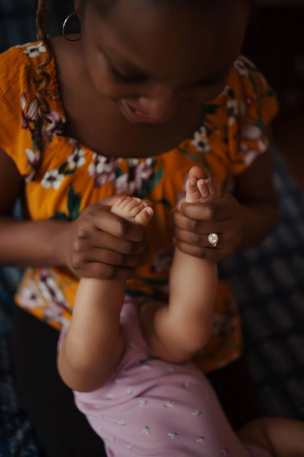 A woman in a floral orange dress holds a child's hands and helps the child stretch their arms. The woman wears a ring with a large pearl. The child wears pink pajamas with small white and blue patterns.