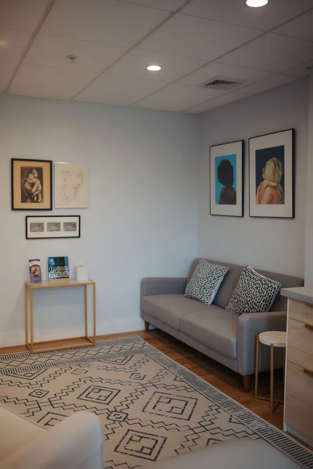 Living room corner with artwork on the wall, gray sofa with patterned cushions, a small side table, and a black and white geometric rug.