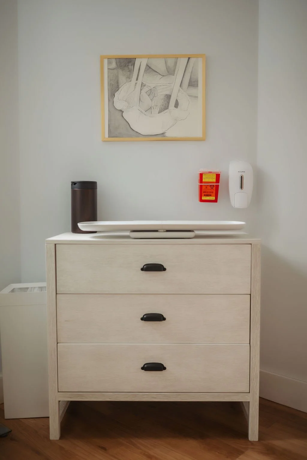 A white changing table with black handles, positioned against a light gray wall. On top of the table is a white changing pad. To the left of the pad is a black container. On the wall above, there's a framed drawing or photograph. To the right of the 