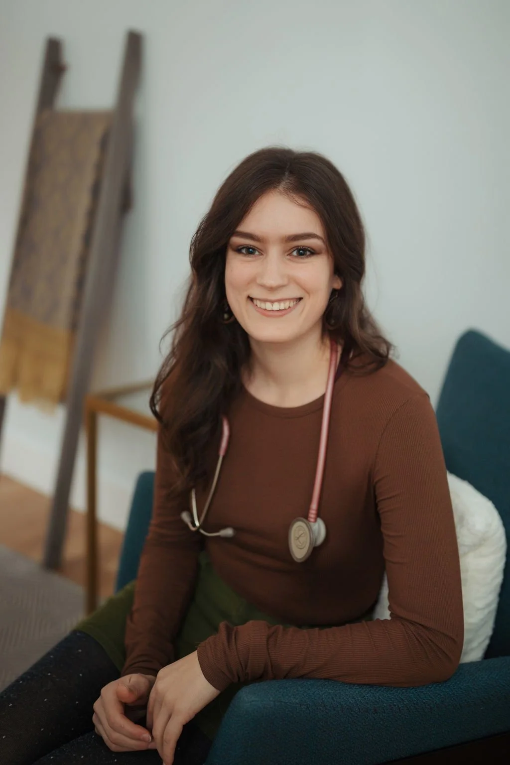 A young woman with long brown hair, smiling, sitting on a blue chair indoors. She is wearing a brown long-sleeve shirt and a stethoscope around her neck.