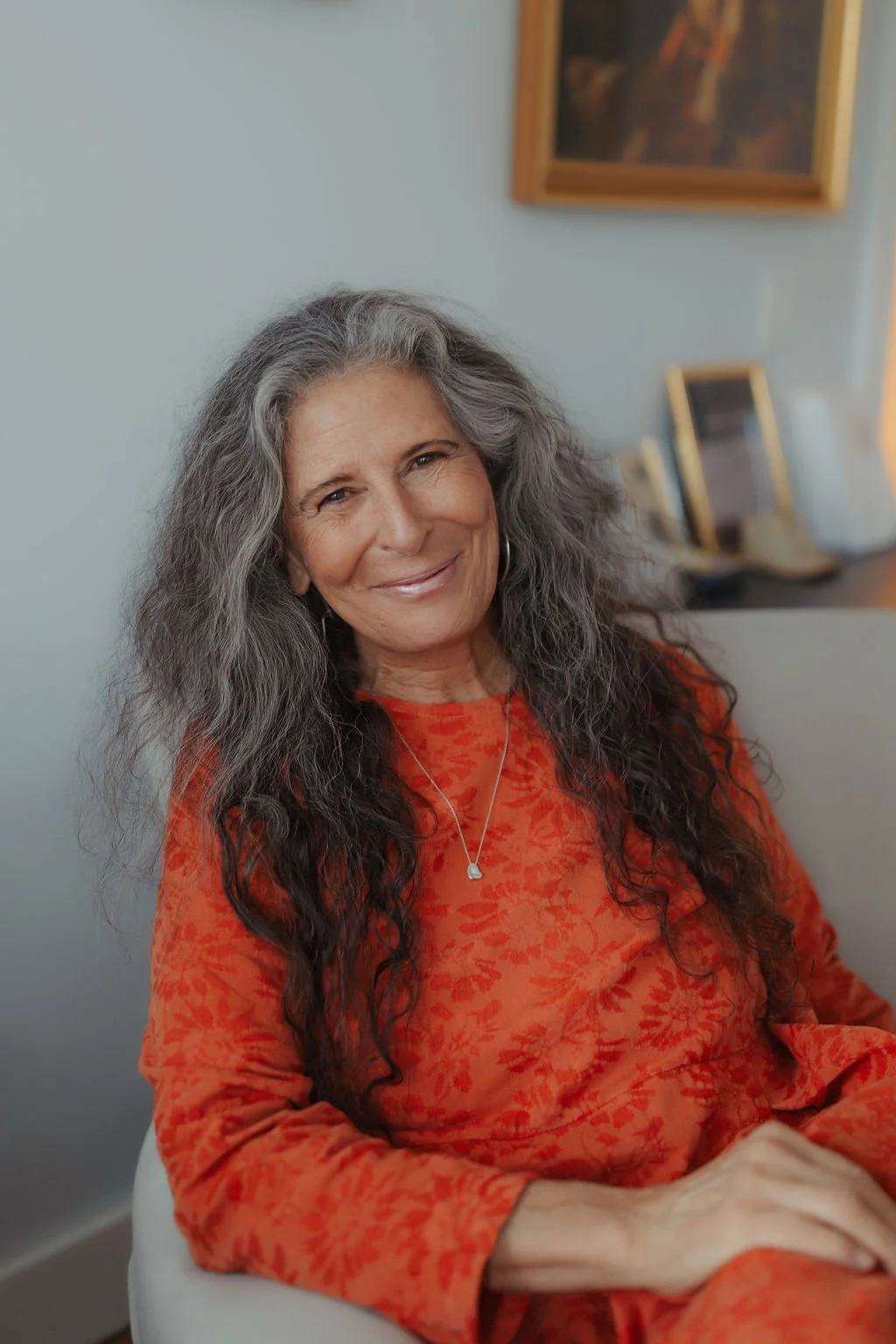 A woman with long, curly gray hair, smiling, wearing an orange floral dress and a silver necklace, sitting indoors near a wall with framed pictures.