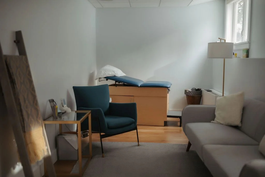 A hospital room with a blue armchair, a gray sofa, a bedside table, a lamp, and a hospital bed with a pillow, near a window letting in natural light.