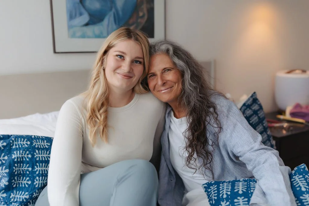 A young woman with blonde hair and an older woman with long gray hair sitting close together on a bed, smiling at the camera. The bed has blue and white patterned pillows, and the room has a framed picture and a bedside table with a lamp.