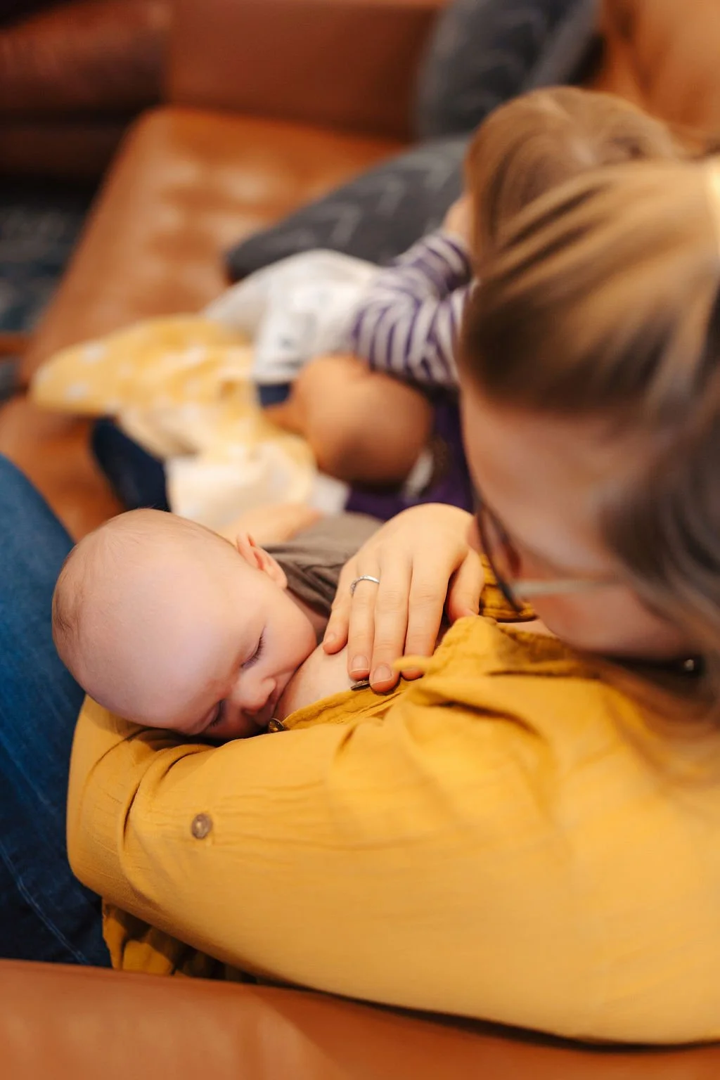 A woman holding a sleeping baby on her chest, with a toddler sitting next to them on a brown leather couch.