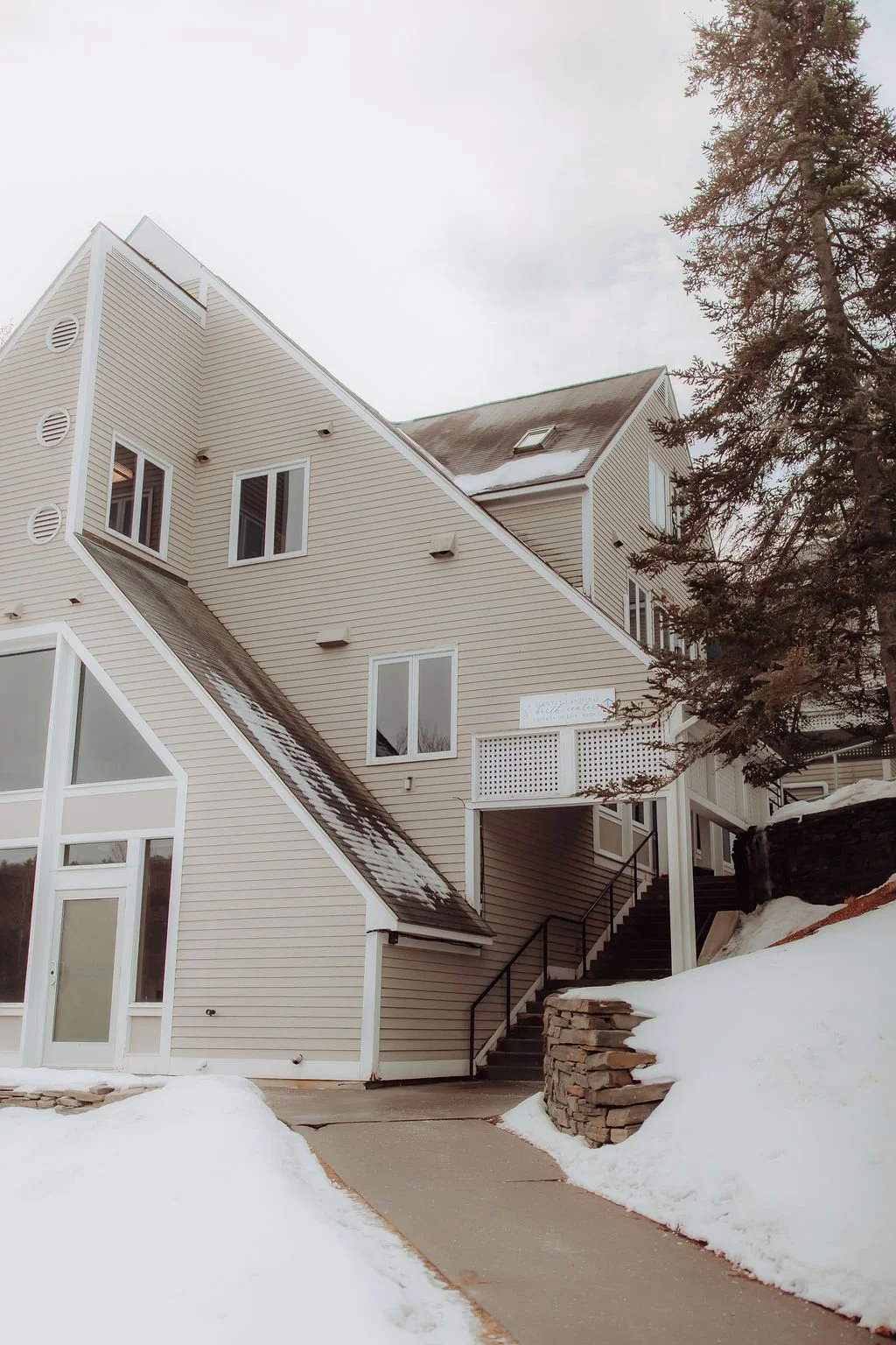 A multi-story house with beige siding and large windows, set in a snowy landscape with a paved path and a stone retaining wall, partially covered by snow, under a cloudy sky.