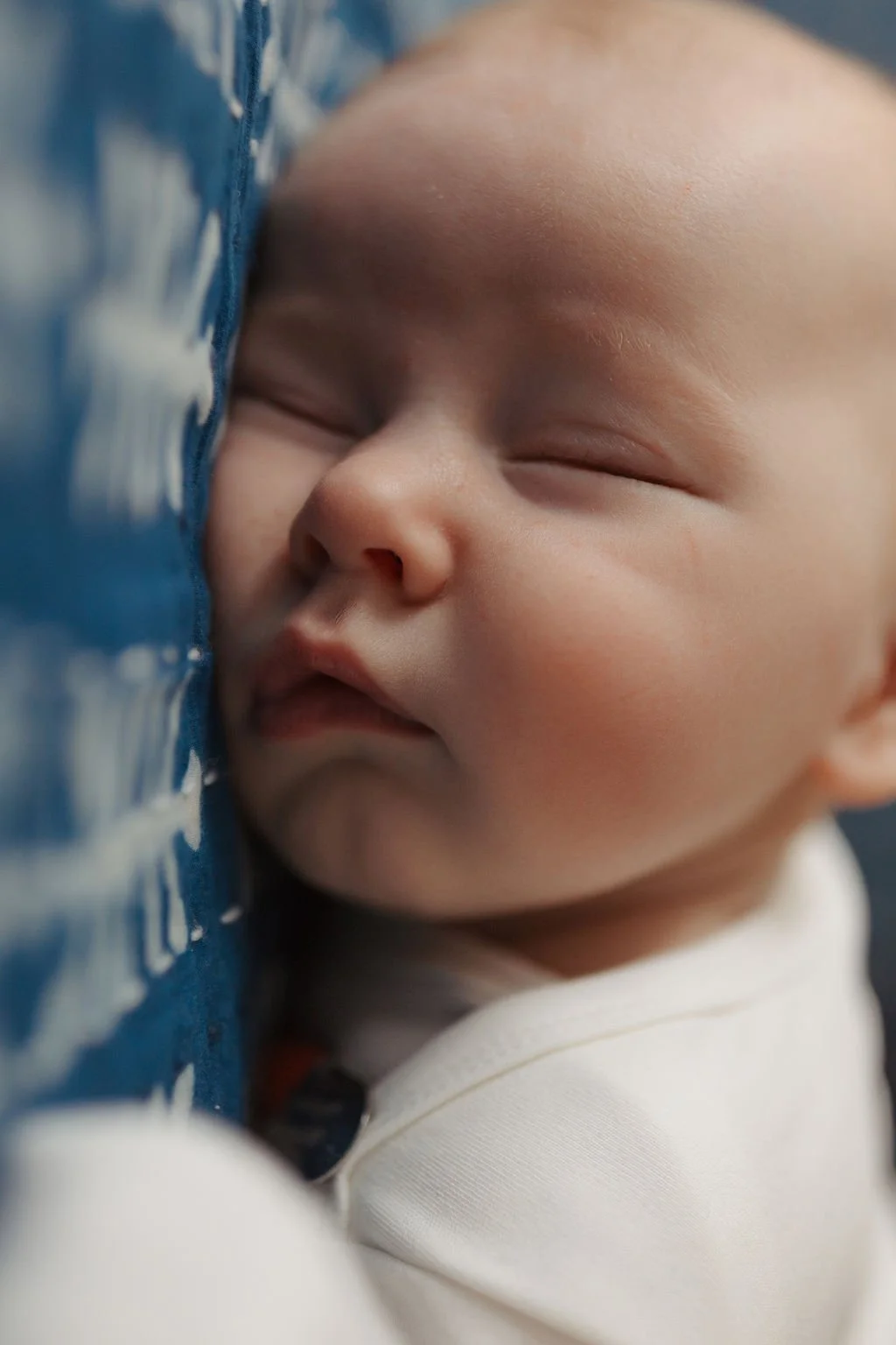 Close-up of a sleeping baby resting peacefully with eyes closed, face relaxed, and soft skin, lying against a blue and white patterned fabric.