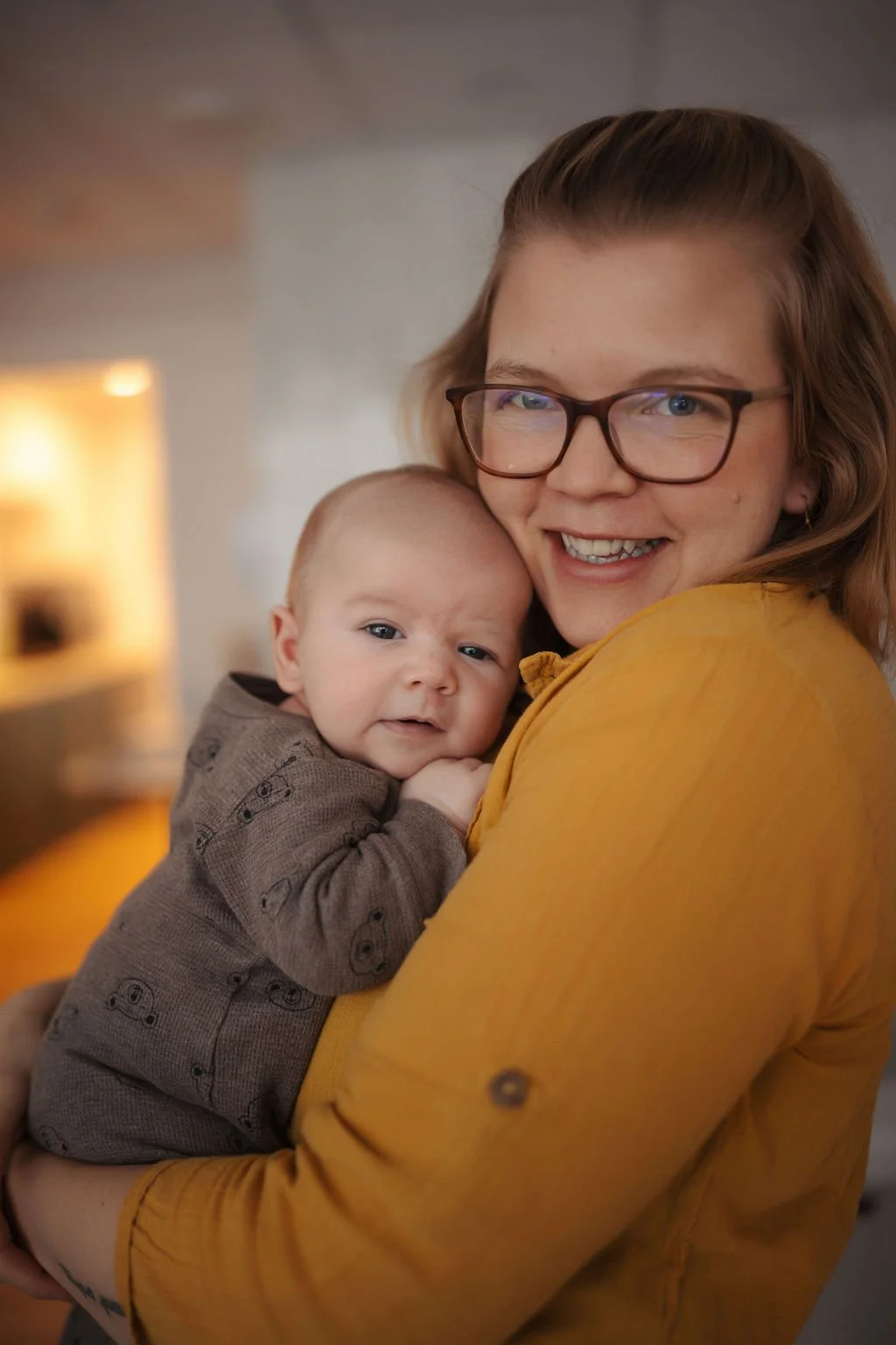 A woman with glasses and a yellow shirt smiling while holding a baby with brown hair and a brown onesie.
