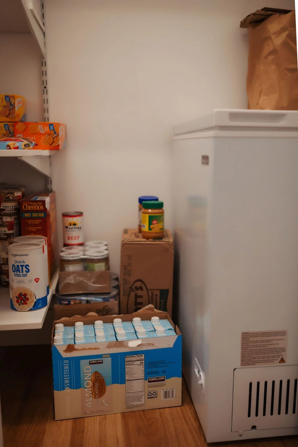 A pantry corner with shelves stocked with cereal and food boxes, jars, and canned goods, next to a white freezer and a brown paper bag on top.