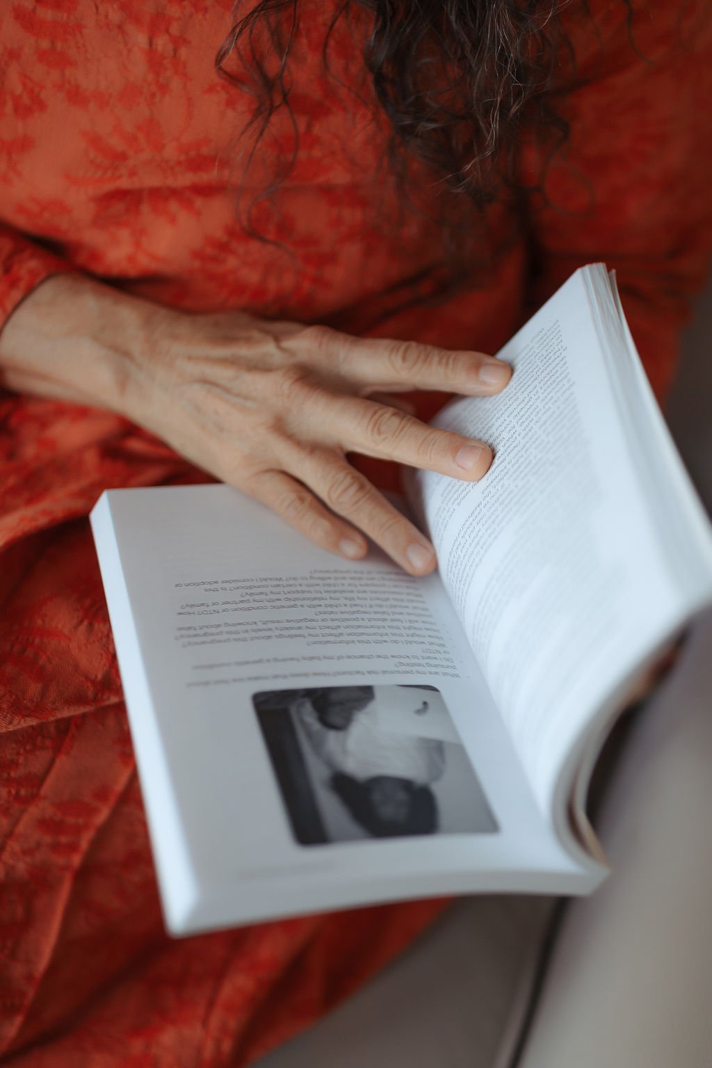 Close-up of an elderly person with visible wrinkles and aged skin, wearing a red patterned dress with a floral design, reading a booklet or magazine with black and white photos.