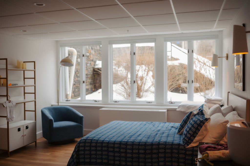 Cozy bedroom with a large window showing snowy outdoor scene, bed with blue patterned bedding, blue armchair, and modern lighting fixtures.