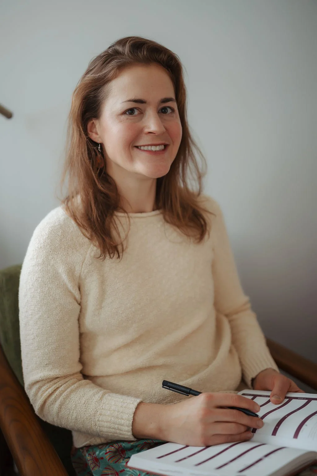 A woman with shoulder-length brown hair, smiling, sitting on a green armchair, holding a pen and a notepad with black lines, dressed in a cream sweater and patterned pants, against a plain gray background.