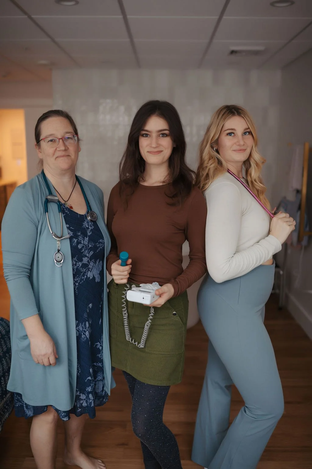 Three women, including two healthcare professionals and one patient, standing together in a room with a plain wall and wooden floor.