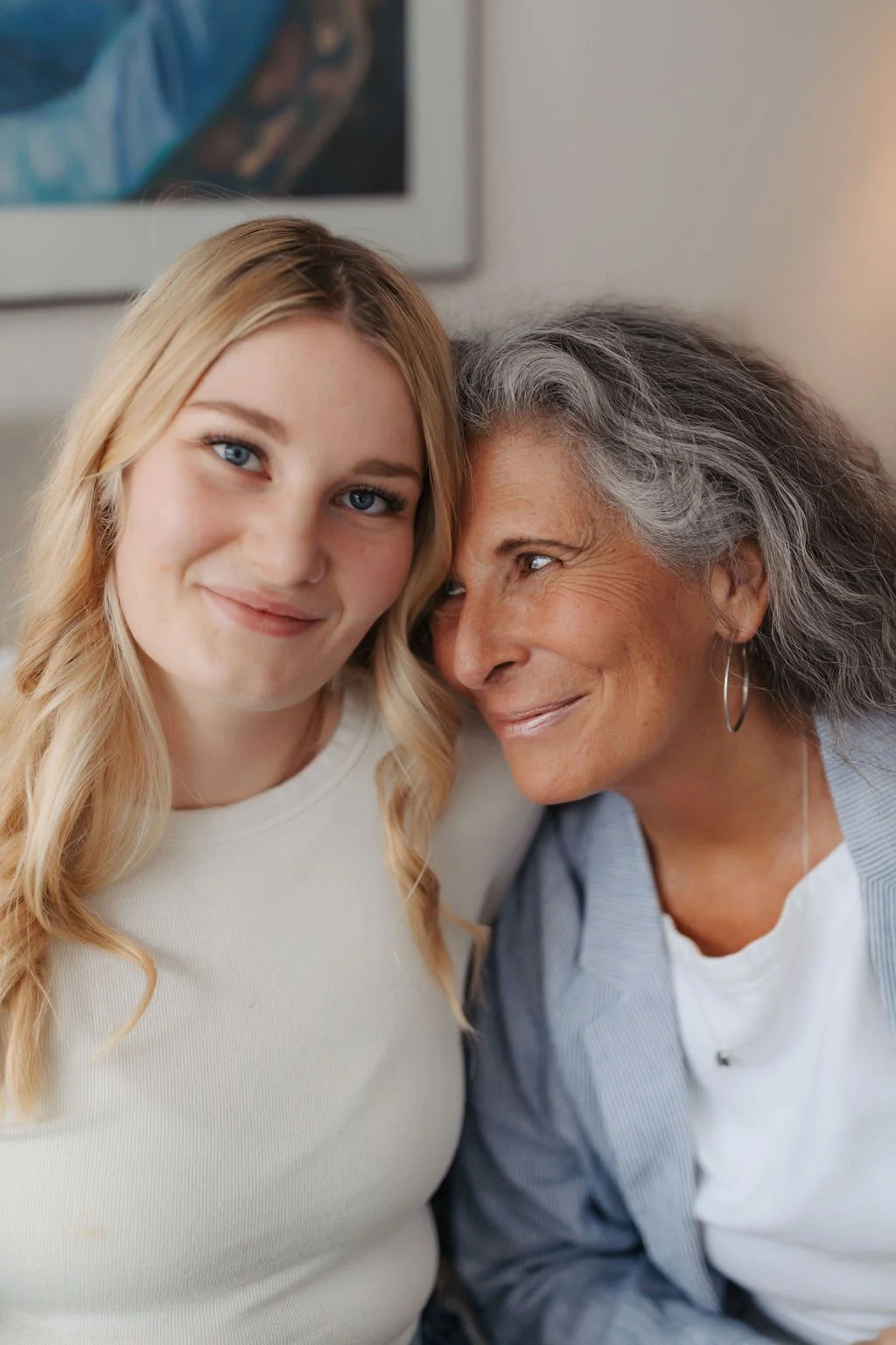 A young woman with blonde hair and a senior woman with gray hair smiling close together indoors.