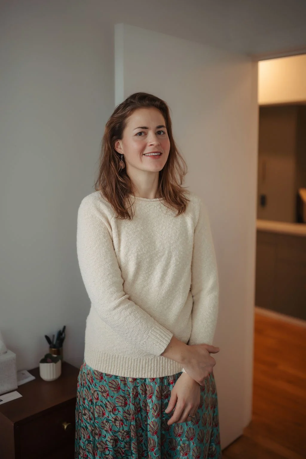 A woman with shoulder-length brown hair wearing a cream sweater and a patterned skirt standing indoors near a wall, with a small table and some office supplies visible in the background.