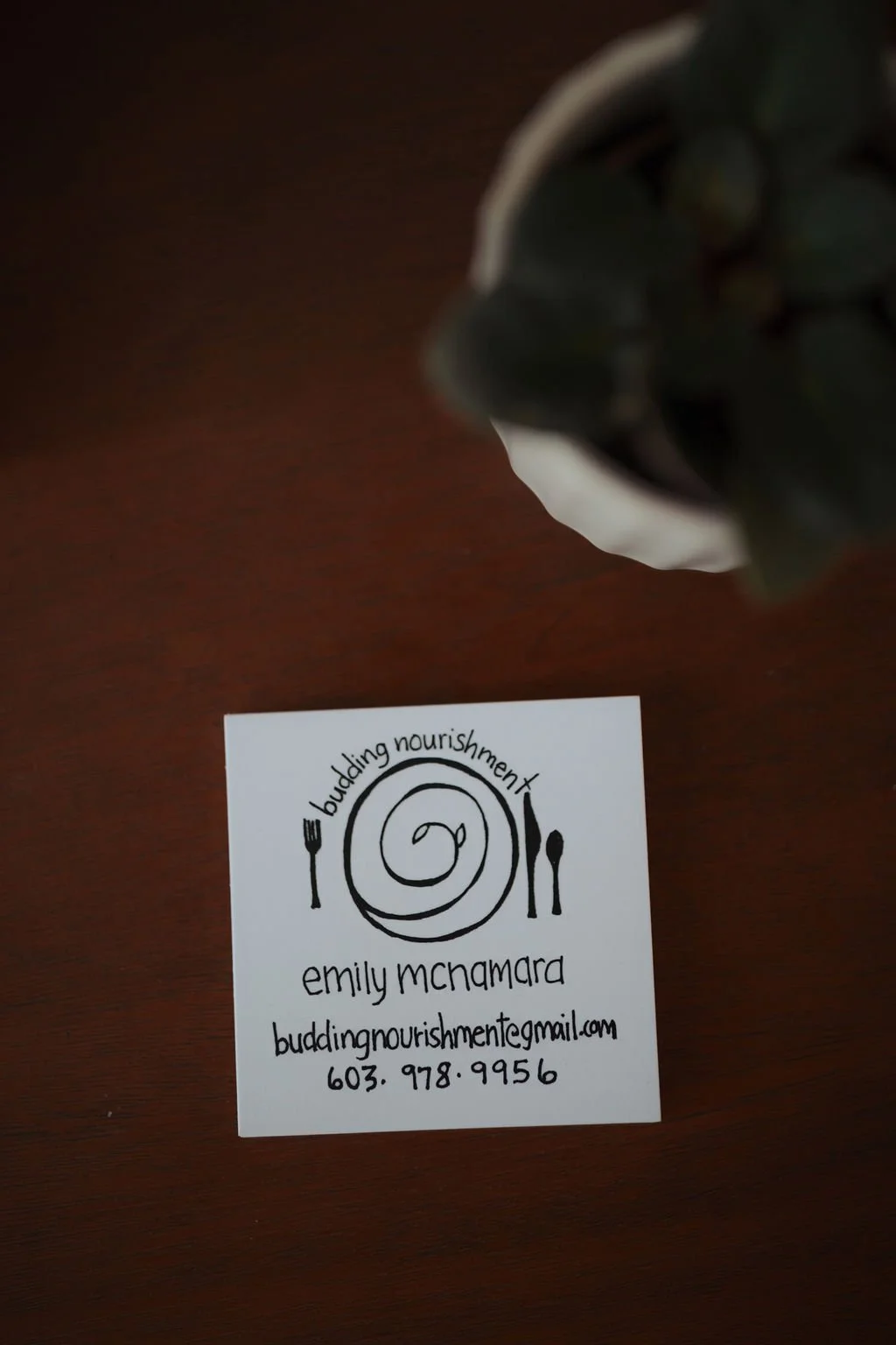 A business card for Emily McNamara's 'Budding Nourishment' features a black and white design with a spiral plate, fork, and knife, and contact information on a wooden surface, with part of a food takeout container visible at the top edge.