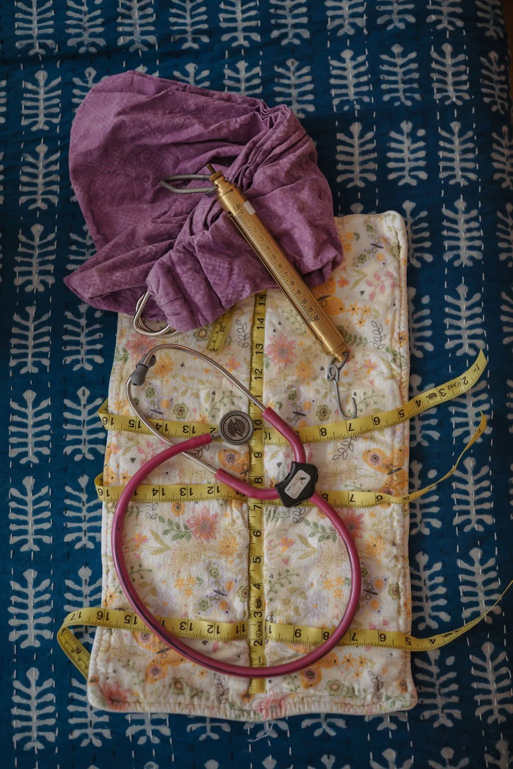 Medical items including a pink stethoscope, a yellow measuring tape, a blood pressure cuff, and a surgical mask on a floral cloth, with a purple surgical gown and a thermometer on a blue patterned fabric background.